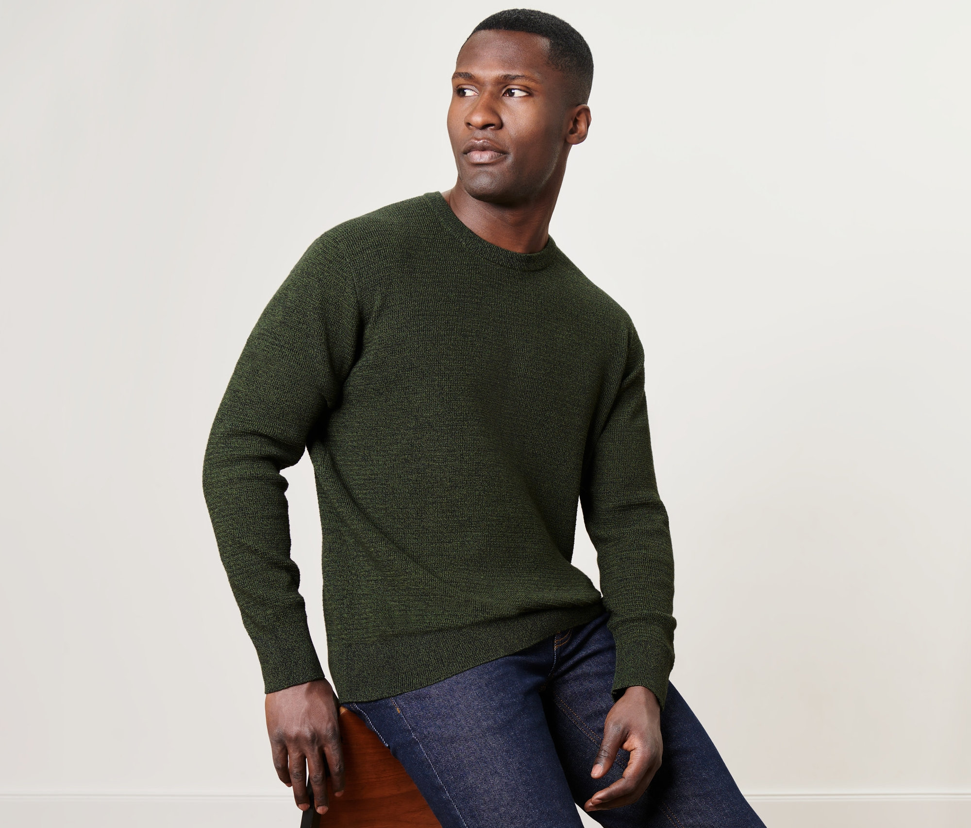 Adam sandalyede oturuyor ve haki rengi, yuvarlak yakalı bir kazak giyiyor.