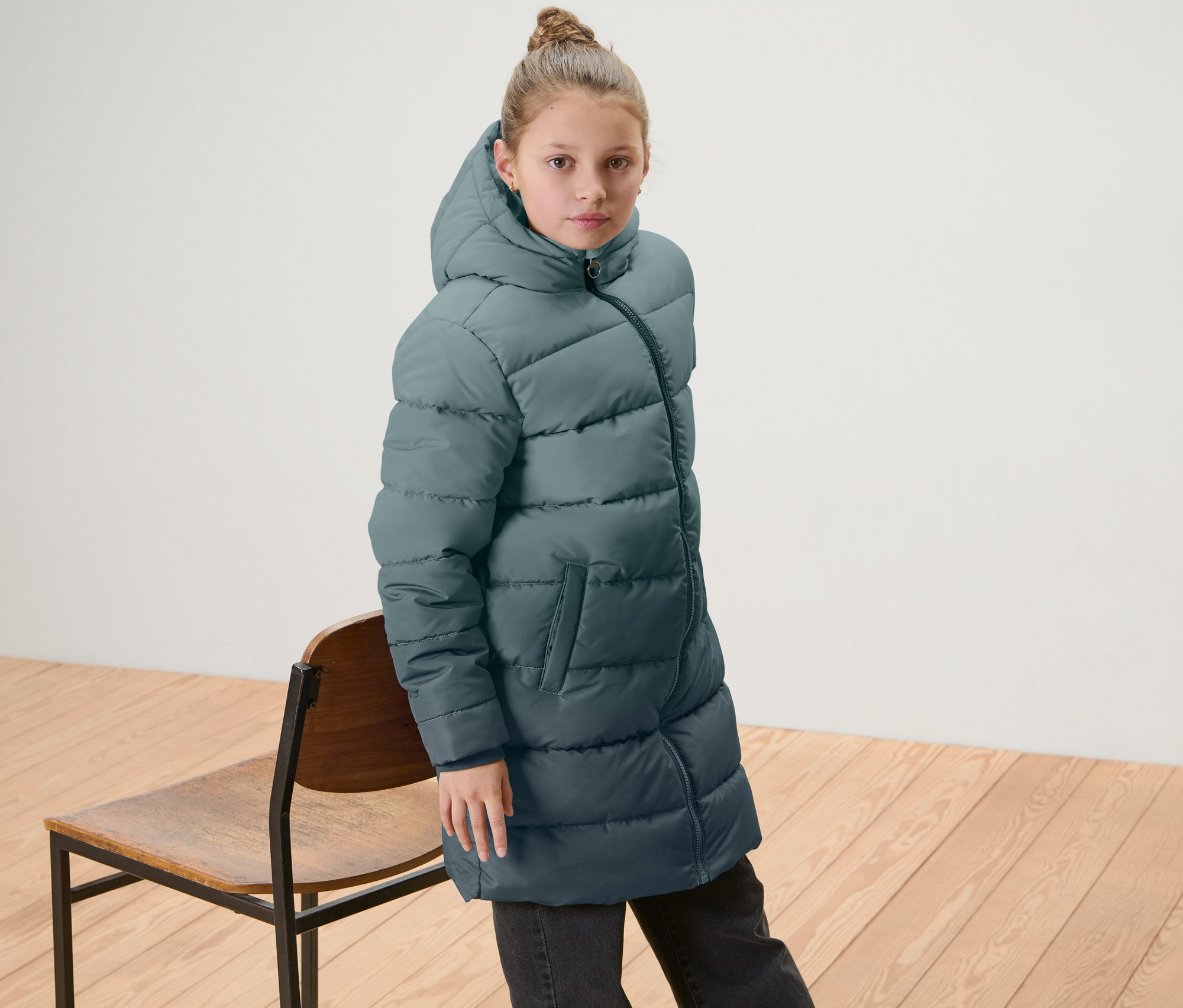 Kız çocuğu, renk geçişli çocuk termal paltosunu giymiş ve ahşap bir sandalyenin yanında duruyor.