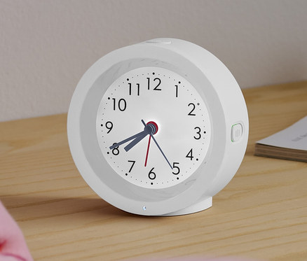 Arkadan aydınlatmalı beyaz analog bir quartz çalar saat, bir yatağın yanındaki komodinin üzerinde duruyor.