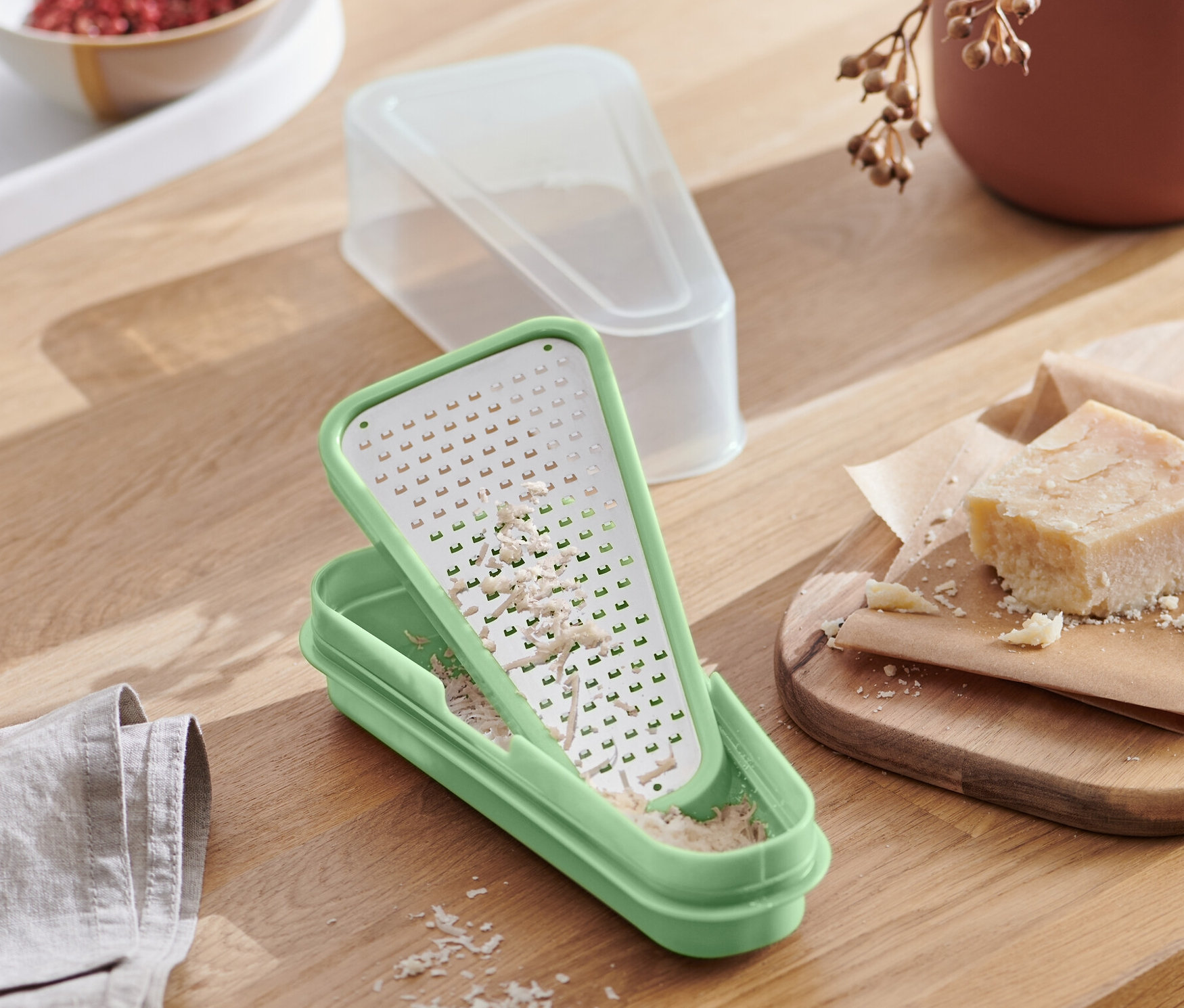 Rendelenmiş peynirle birlikte yeşil renkli 2'si 1 arada parmesan rendesi, bir tabak makarnanın yanında ahşap bir masa üzerinde duruyor.