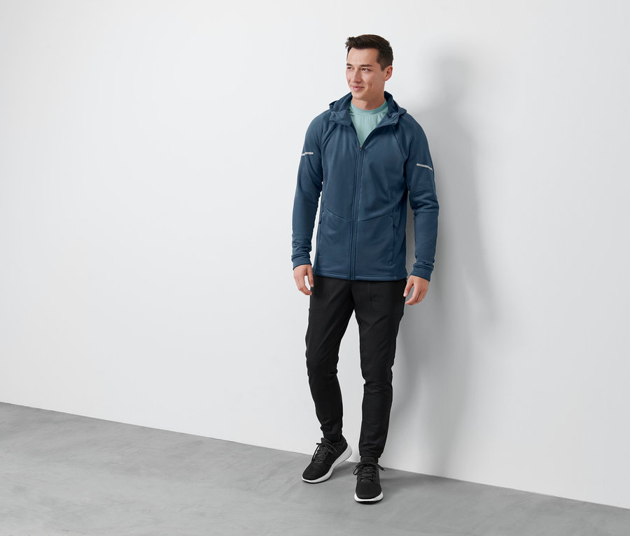 Bir adam ayakta duruyor ve mavi bir termal ceket, siyah bir termal ceket, fonksiyonel bir termal pantolon ve siyah ayakkabılar giyiyor.