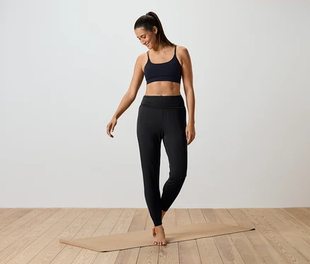 Kadın yoga matının üzerinde duruyor, siyah spor sütyeni ve siyah taytı var.