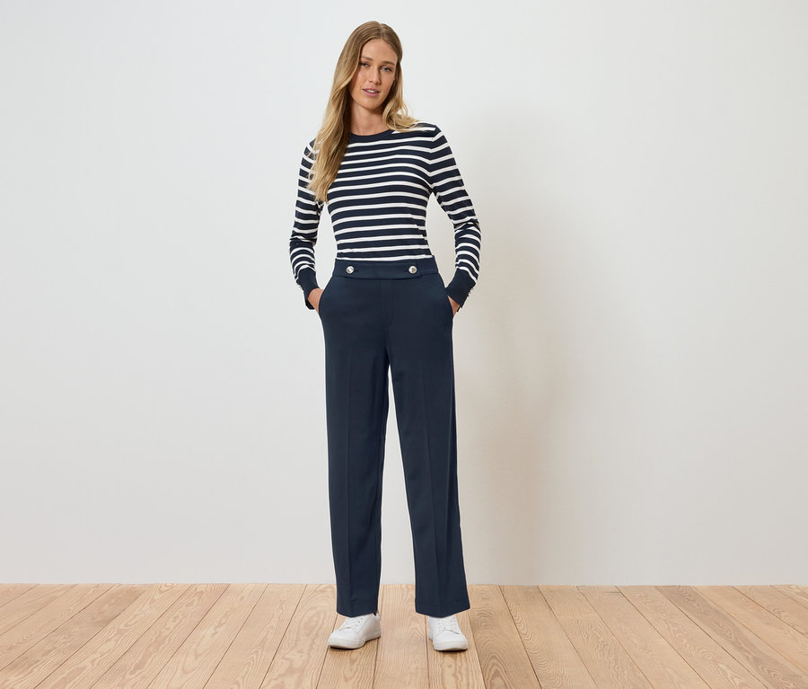 Bir kadın elleri cebinde ayakta duruyor. Çizgili, uzun kollu bir tişört ve lacivert pantolon giyiyor.