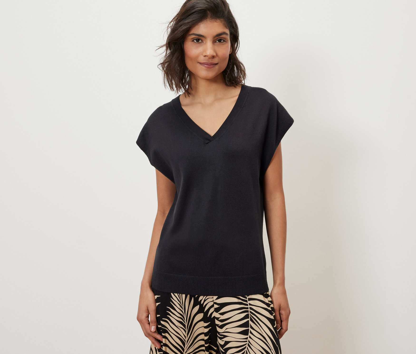 Kadın siyah, kısa kollu, ince örgülü bir kazak ve desenli bir pantolon giyiyor.