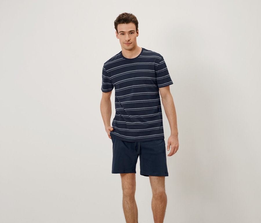 Bir adam ayakta duruyor ve elleri ceplerinde. Yatay çizgili bir tişört ve şort giyiyor.