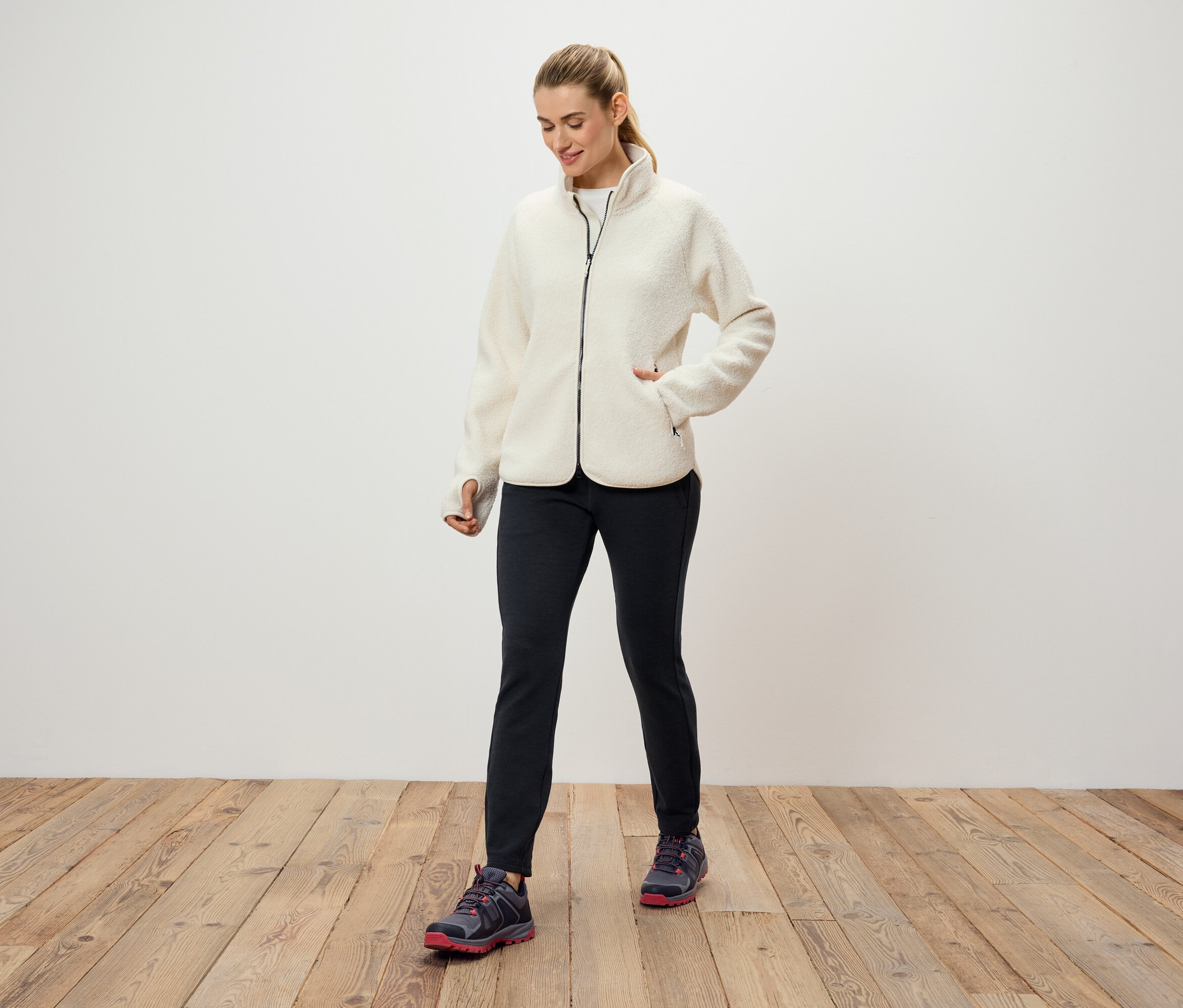 Krem rengi polar ceket, siyah pantolon ve spor ayakkabı giyen bir kadın, beyaz bir duvarın önünde ahşap bir zeminde duruyor.