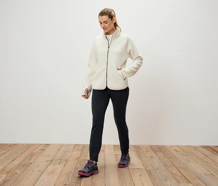 Krem rengi polar ceket, siyah pantolon ve spor ayakkabı giyen bir kadın, beyaz bir duvarın önünde ahşap bir zeminde duruyor.