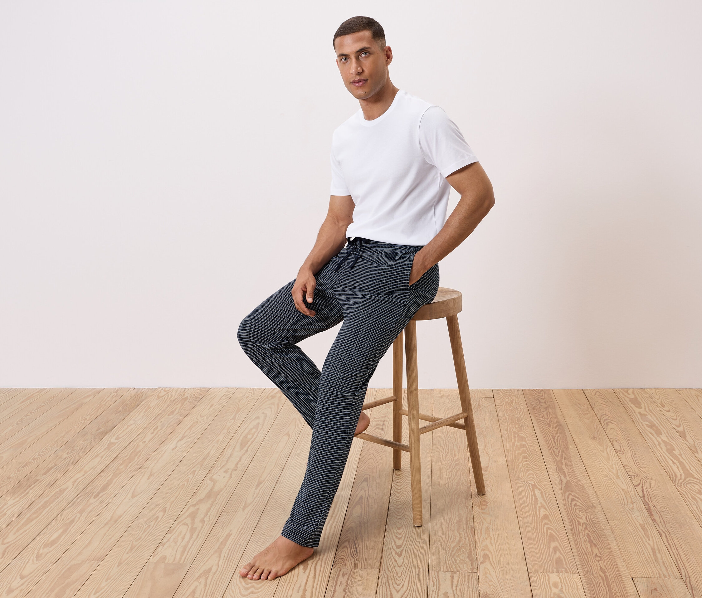 Adam bir taburede oturuyor, beyaz bir tişört ve desenli pantolon giyiyor.