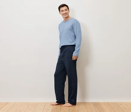 Bir adam beyaz bir duvarın önünde çıplak ayakla duruyor, açık mavi bir kazak ve üzerinde puantiyeler olan lacivert bir pijama altı giyiyor.