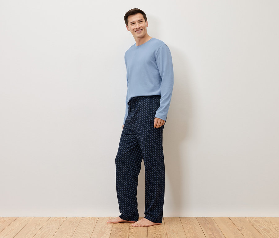 Bir adam beyaz bir duvarın önünde çıplak ayakla duruyor, açık mavi bir kazak ve üzerinde puantiyeler olan lacivert bir pijama altı giyiyor.