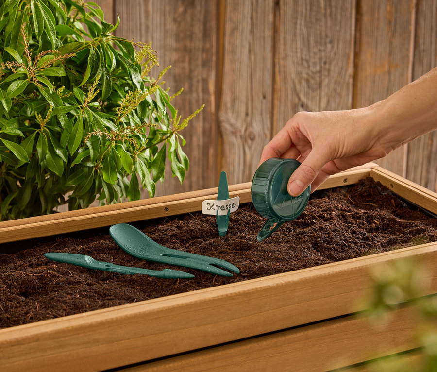 Bir el, içinde yeşil bahçe aletleri ve "Kresse" yazılı bir etiket bulunan toprak dolu ahşap bir kutuya küçük bir yeşil sulama kabından su döküyor.