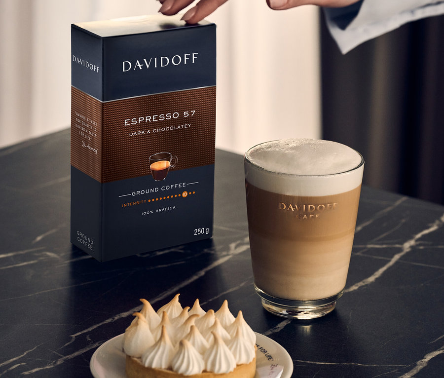 Bir el, DAVIDOFF CAFÉ Espresso 57 250g Gemahlen paketine dokunuyor, yanında bir latte macchiato ve tartölet var.