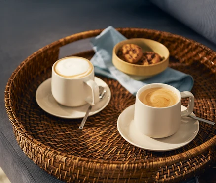 Biri süt köpüklü, diğeri latte art desenli iki açık renk kahve fincanı, kaşıklarıyla birlikte uyumlu tabaklarda yuvarlak hasır bir tepside duruyor. Tepside ayrıca küçük bir kurabiye kasesi ve açık mavi bir peçete bulunuyor.
