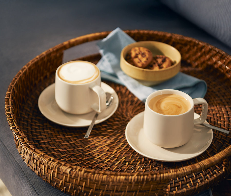 Biri süt köpüklü, diğeri latte art desenli iki açık renk kahve fincanı, kaşıklarıyla birlikte uyumlu tabaklarda yuvarlak hasır bir tepside duruyor. Tepside ayrıca küçük bir kurabiye kasesi ve açık mavi bir peçete bulunuyor.