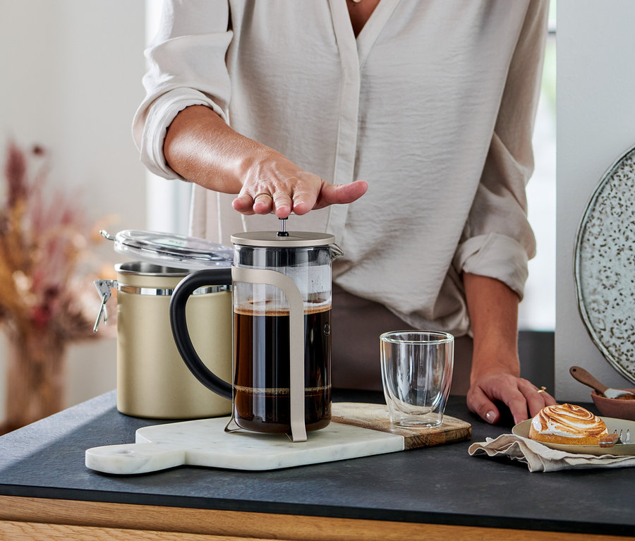 Bir kişi, şampanya rengi 800 ml'lik French Press'in (6 fincan) pistonunu kahveyle dolu olarak aşağı bastırıyor. Yanında bir bardak ve bir pasta var.