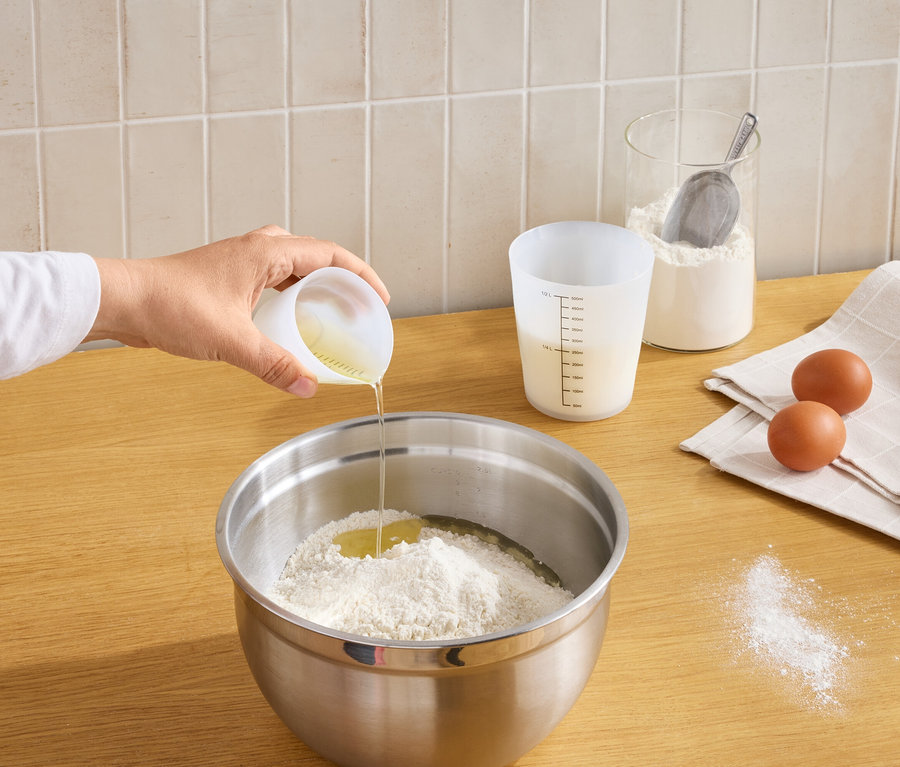 Bir el, küçük bir ölçü kabından un ve kırık yumurta içeren bir kaseye sıvı döküyor. Tezgahta başka bir ölçü kabı, un kavanozu, ekose bir bez üzerinde iki yumurta ve dökülmüş un var.
