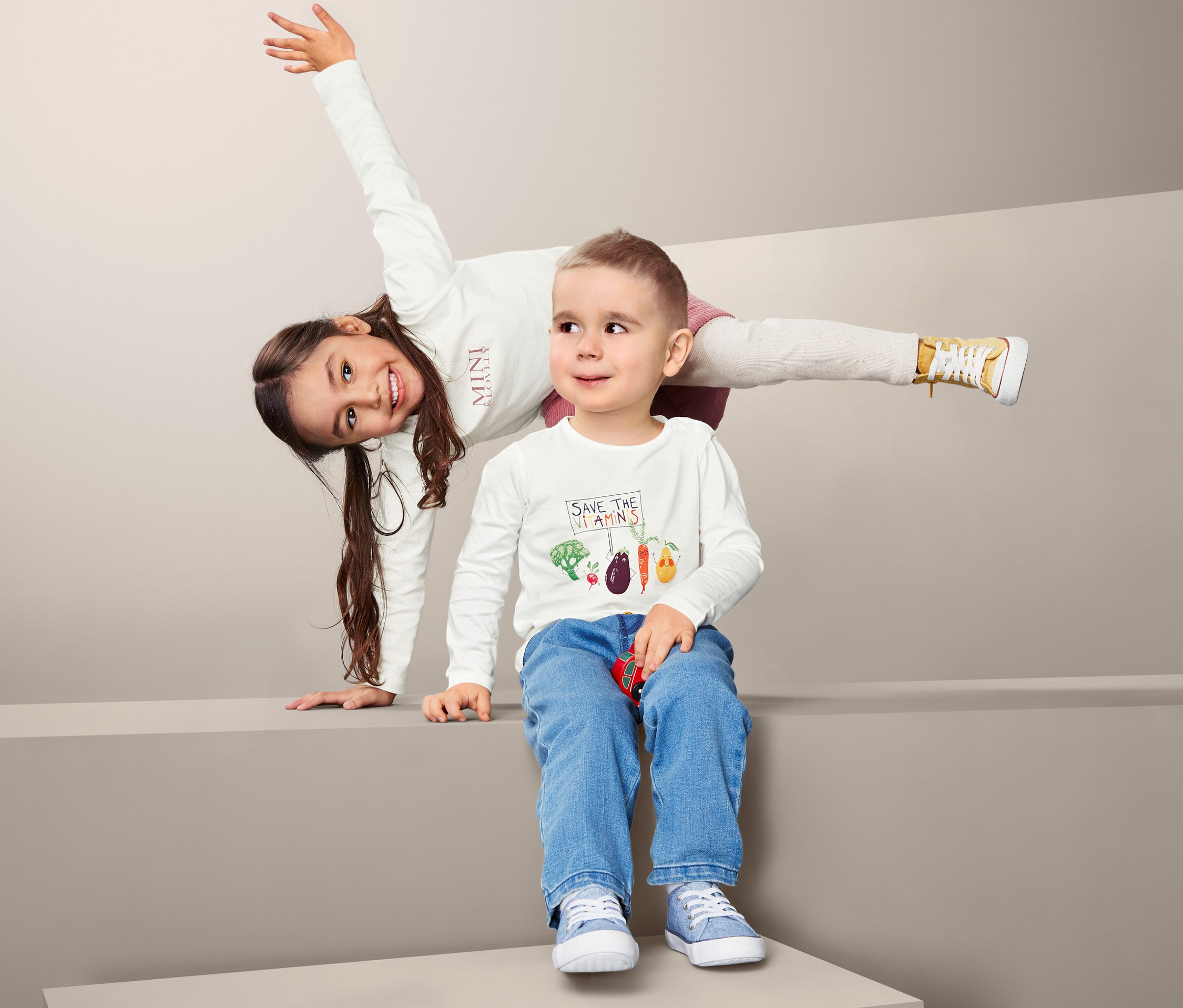 İki çocuk gri bir arka planda poz veriyor. Uzun kollu tişört giyen kız ellerinin üzerinde duruyor, erkek çocuk kot pantolonla oturuyor.