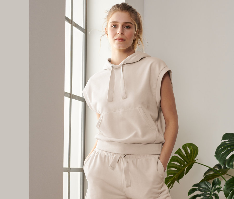 Kadın, kolsuz kapüşonlu spor üstü ve şortla pencere kenarında poz veriyor.