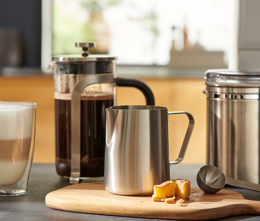 Bir bardak kahve, ahşap bir kesme tahtası üzerinde bir french press ve küçük bir paslanmaz çelik sütlüğün yanında duruyor.