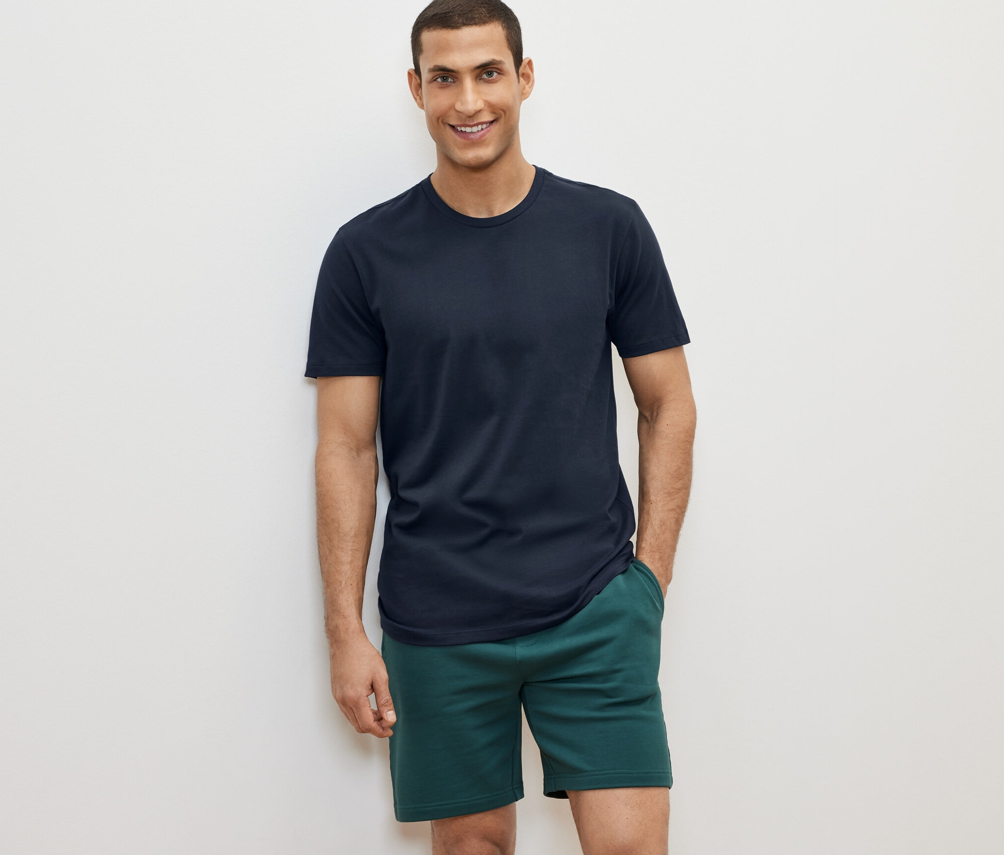 Beyaz bir arka planda, 2 adet bisiklet yakalı tişörtün birini ve yeşil şort giyen bir adam poz veriyor.