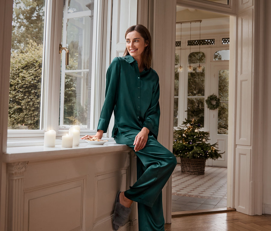 Kadın yeşil ipek pijama üstü, ipek pijama altı ve pelüş terliklerle pencere kenarında oturuyor.