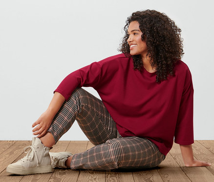 Kadın ahşap zeminde oturuyor, üzerinde bordo renkli 3/4 kollu sweatshirt ve kazayağı desenli jogger pantolon var.