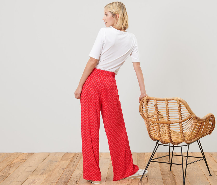 Kadın, rattan bir sandalyenin yanında kırmızı baskılı palazzo pantolon ve beyaz tişörtle poz veriyor.