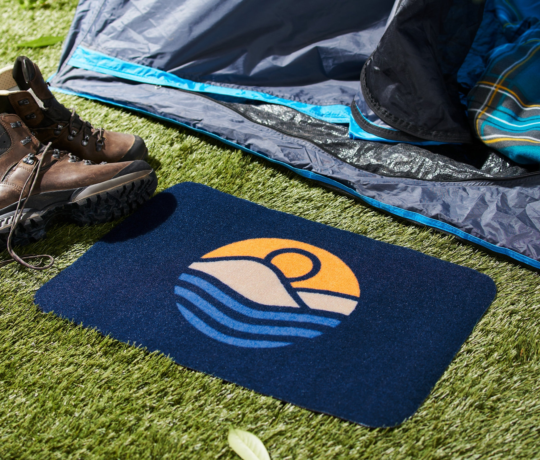 Dağ ve güneş desenli paspas, çimlerin üzerinde bir çadırın önünde yürüyüş botlarının yanında duruyor.