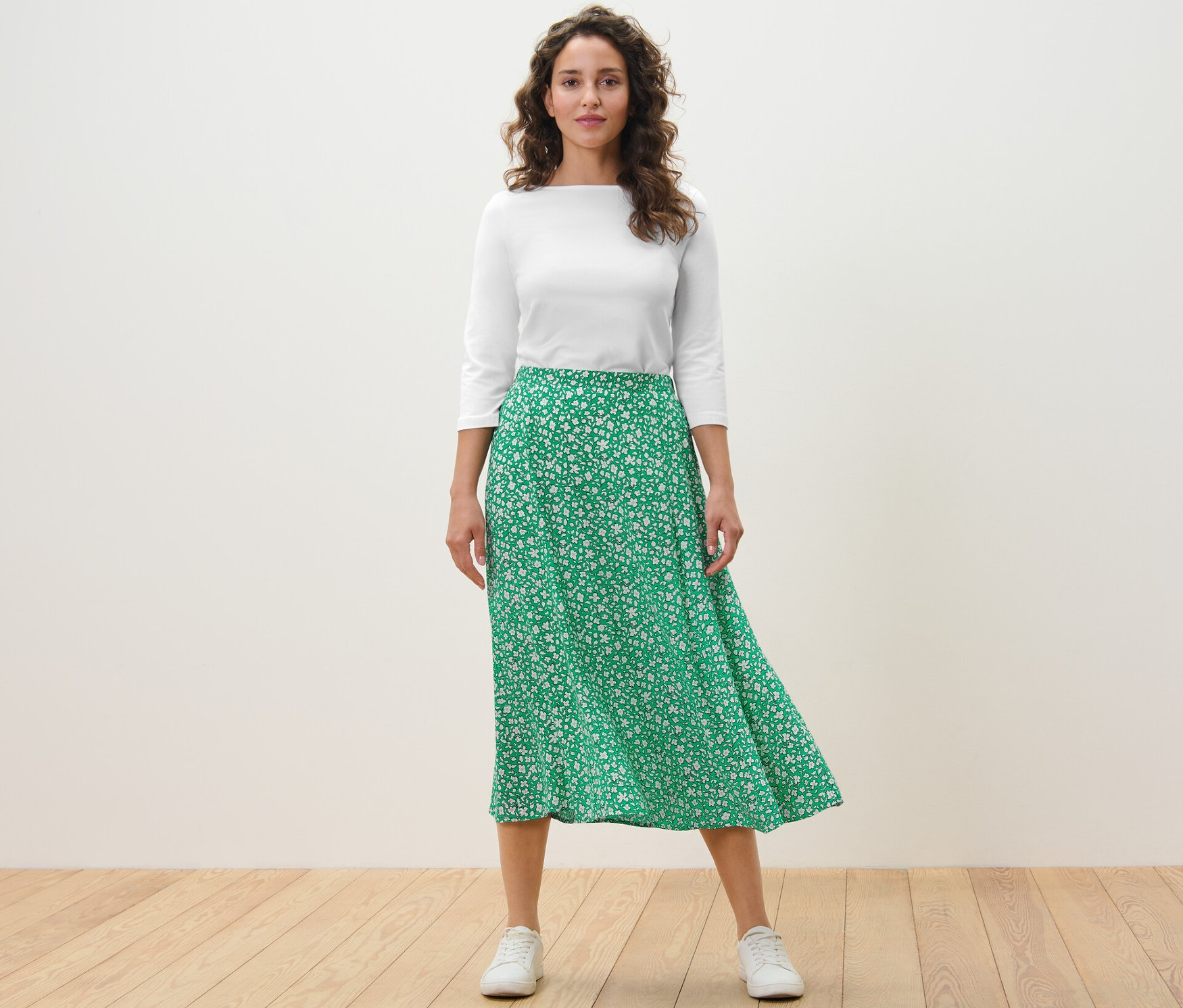 Kadın ayakta duruyor ve Yeşil baskılı midi etek giyiyor.