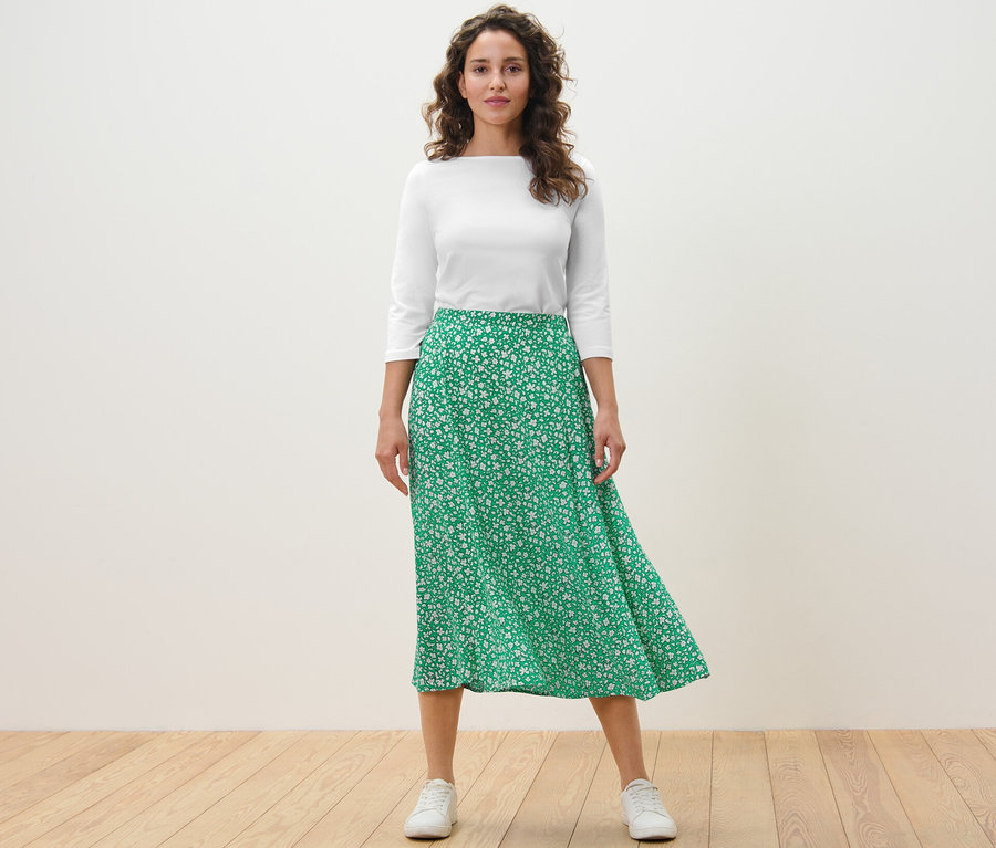 Kadın ayakta duruyor ve Yeşil baskılı midi etek giyiyor.