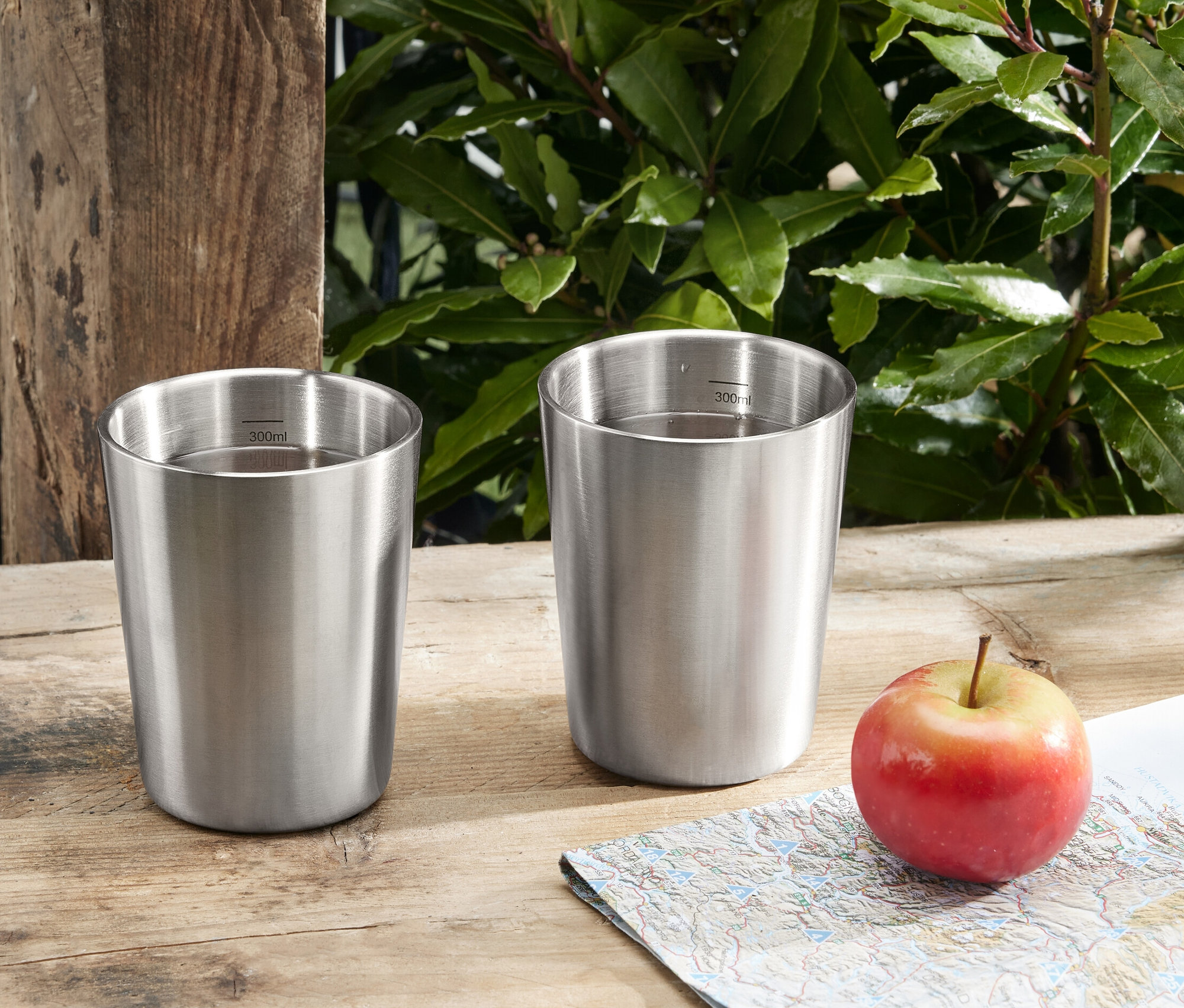 İki adet paslanmaz çelik kamp bardağı, bir elma ve bir haritanın yanında ahşap bir masanın üzerinde duruyor.