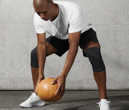 Beyaz tişörtlü ve siyah şortlu adam sağlık topu tutuyor ve 2 Adet Fitness Diz Manşeti takıyor.