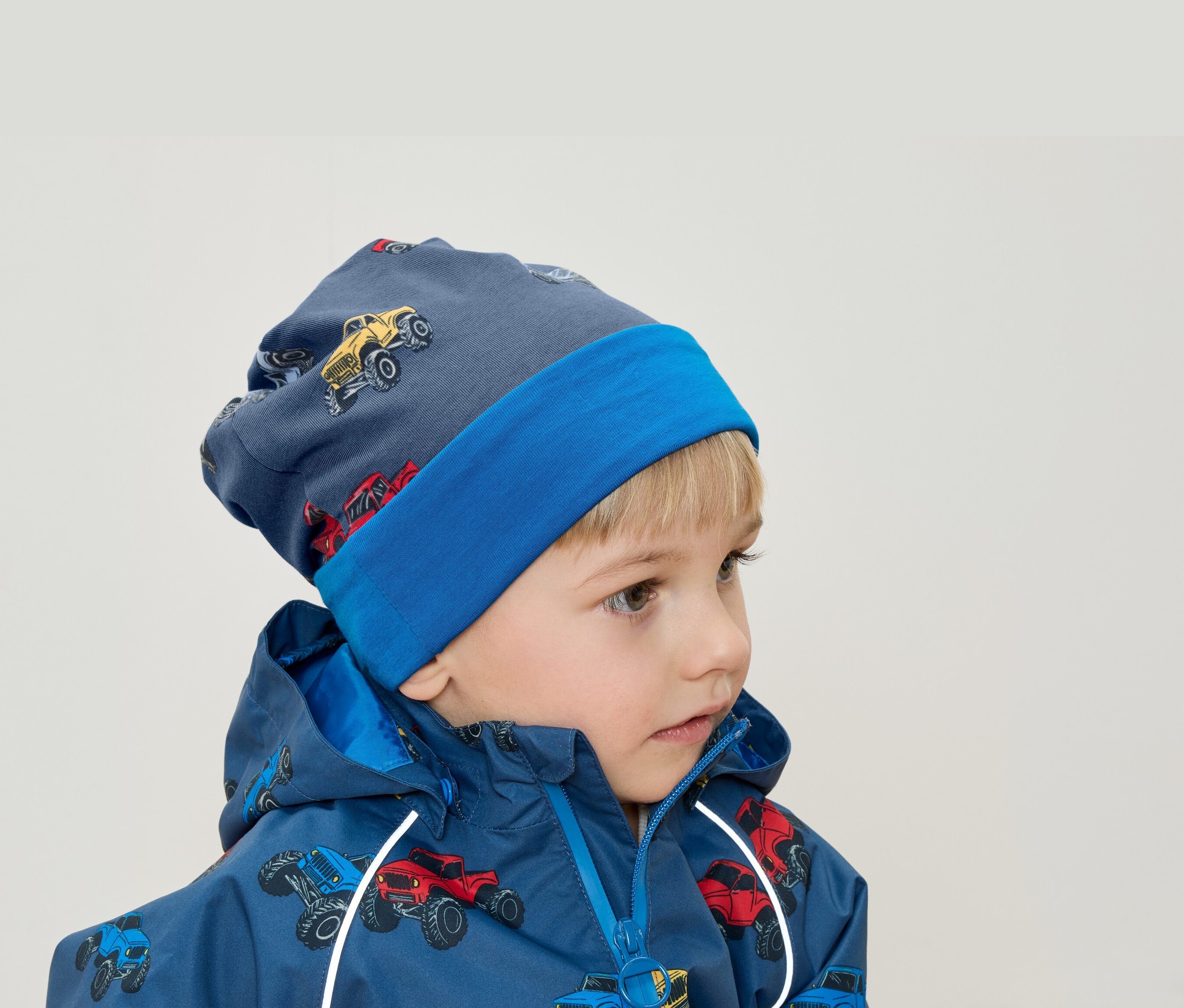 Sarışın saçlı bir çocuk, canavar kamyon baskılı mavi bir şapka ve ceket giyiyor.