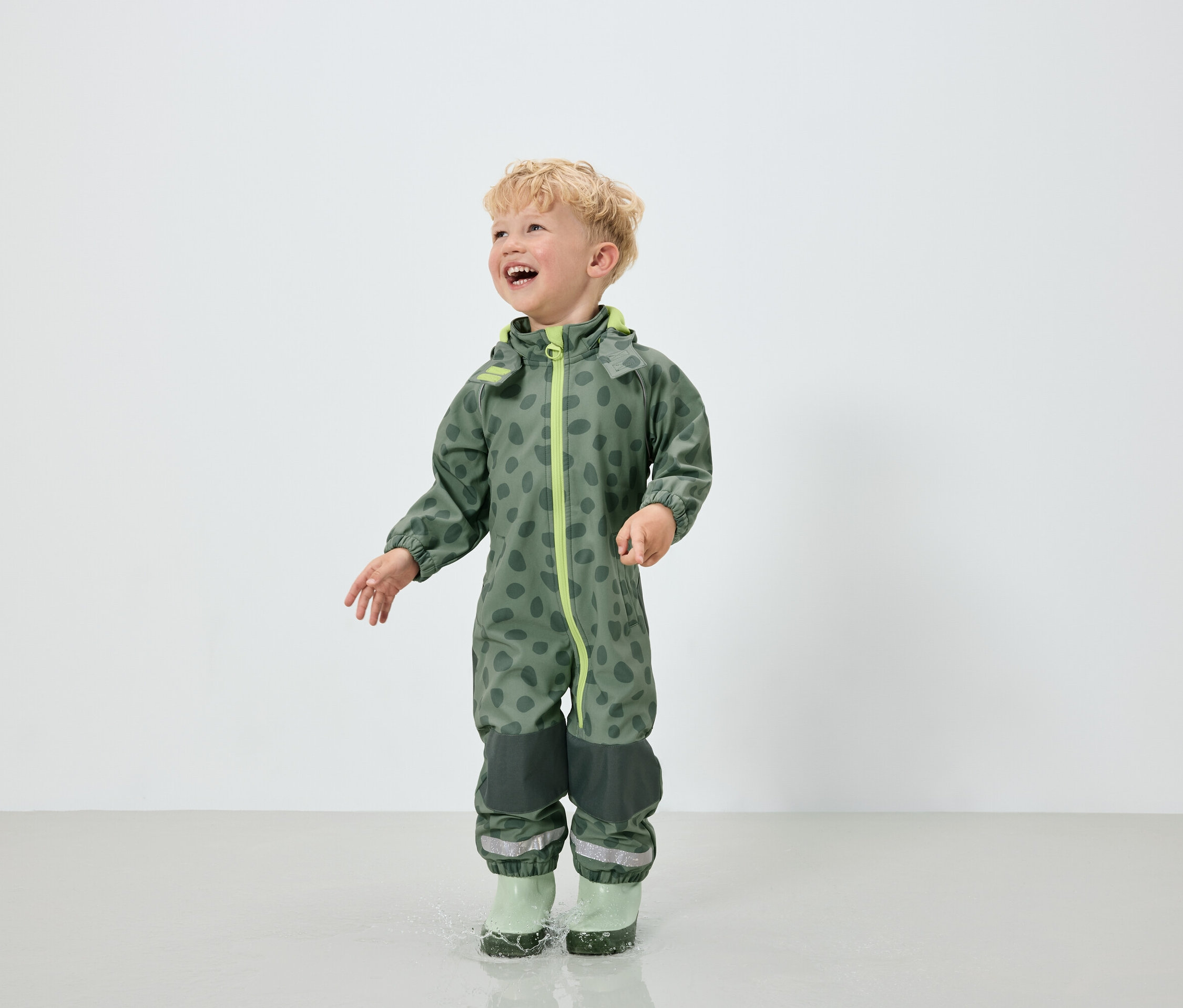 Benekli desenli yeşil tulum ve yeşil lastik çizme giyen bir erkek çocuk su birikintisinin içinde zıplıyor.