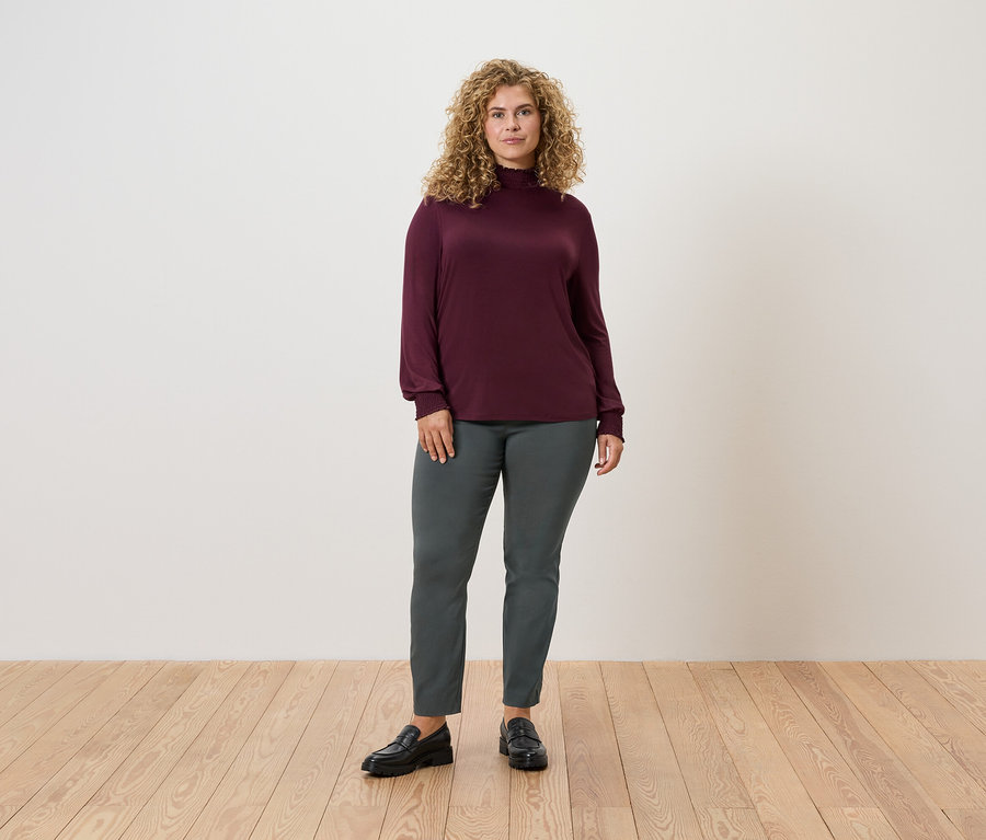 Ahşap zeminde duran bir kadın, koyu yeşil streç pantolon ve bordo uzun kollu bir bluz giyiyor.