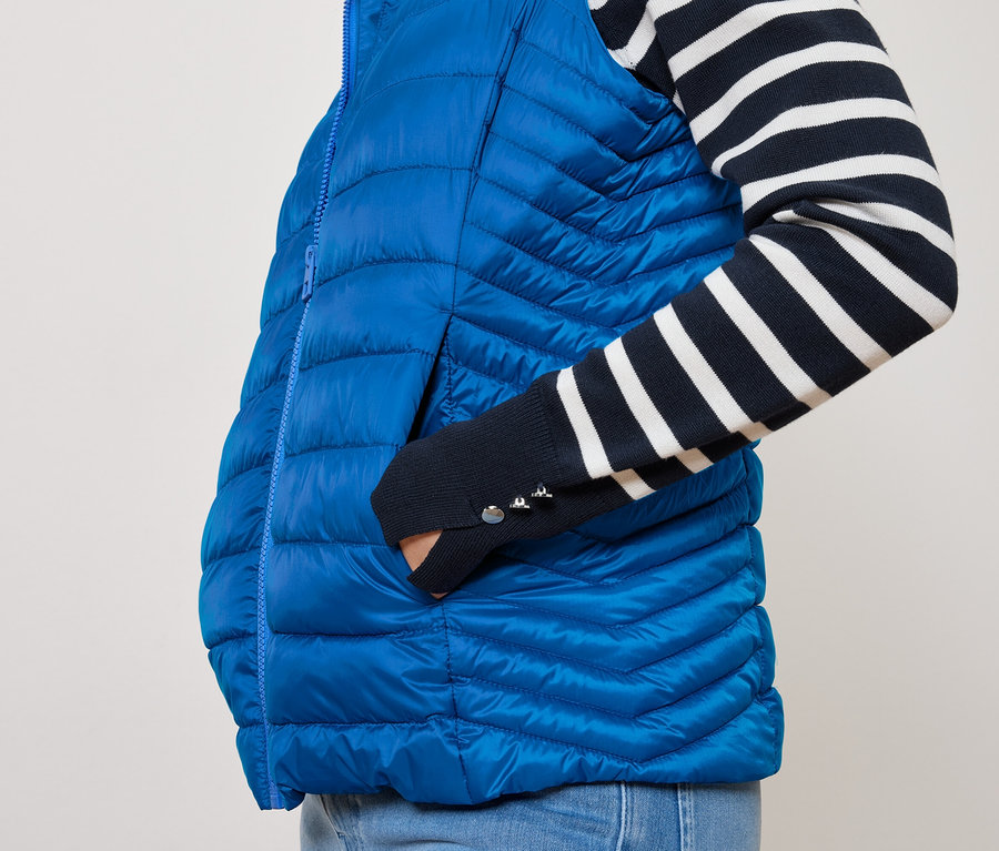 Bir kişi mavi kapitone bir yeleğin cebinde elini tutuyor ve lacivert-beyaz çizgili bir üst giyiyor.