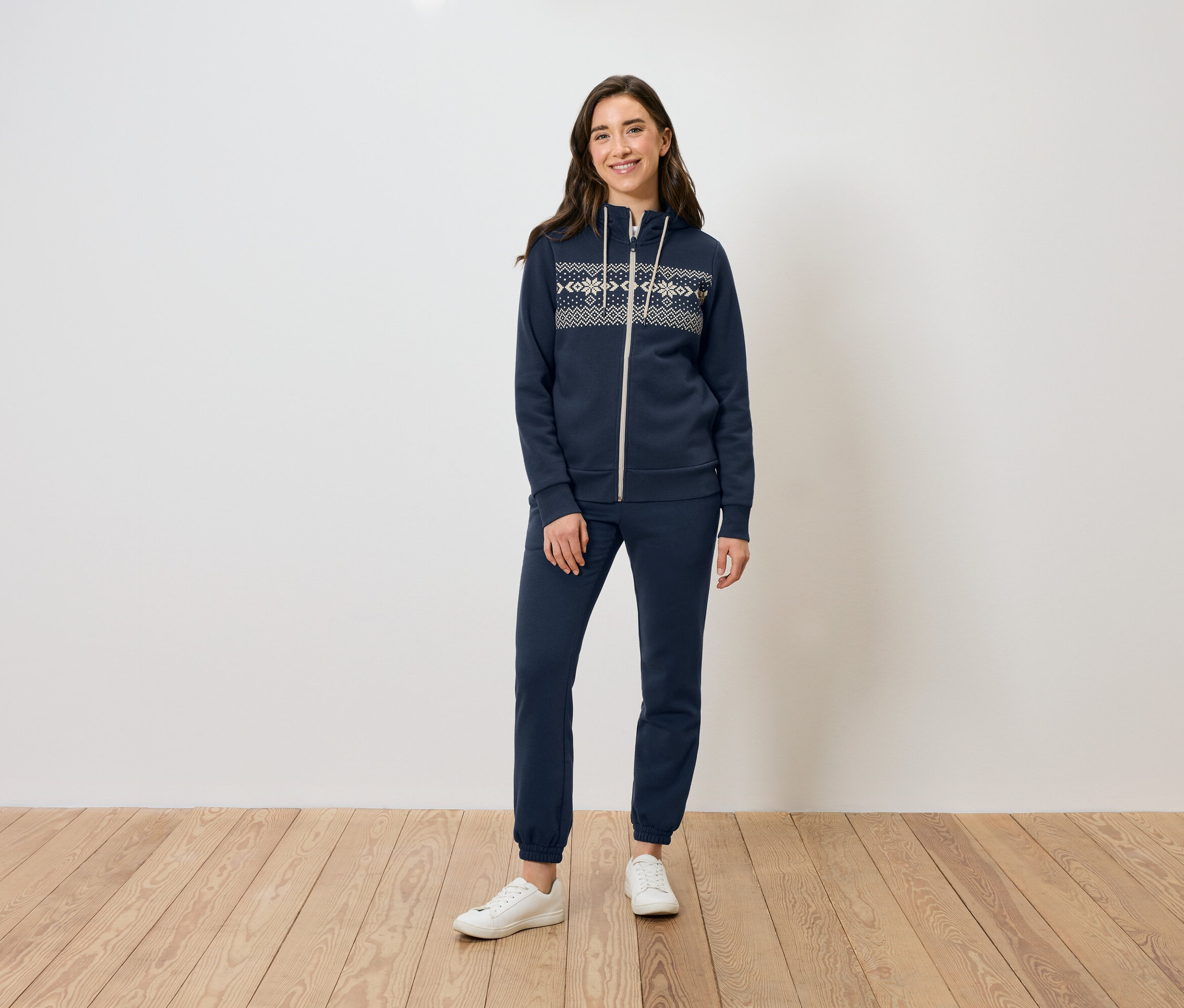 Bir kadın ahşap zeminde duruyor, lacivert fermuarlı kapüşonlu sweatshirt, eşofman altı ve beyaz spor ayakkabı giyiyor.