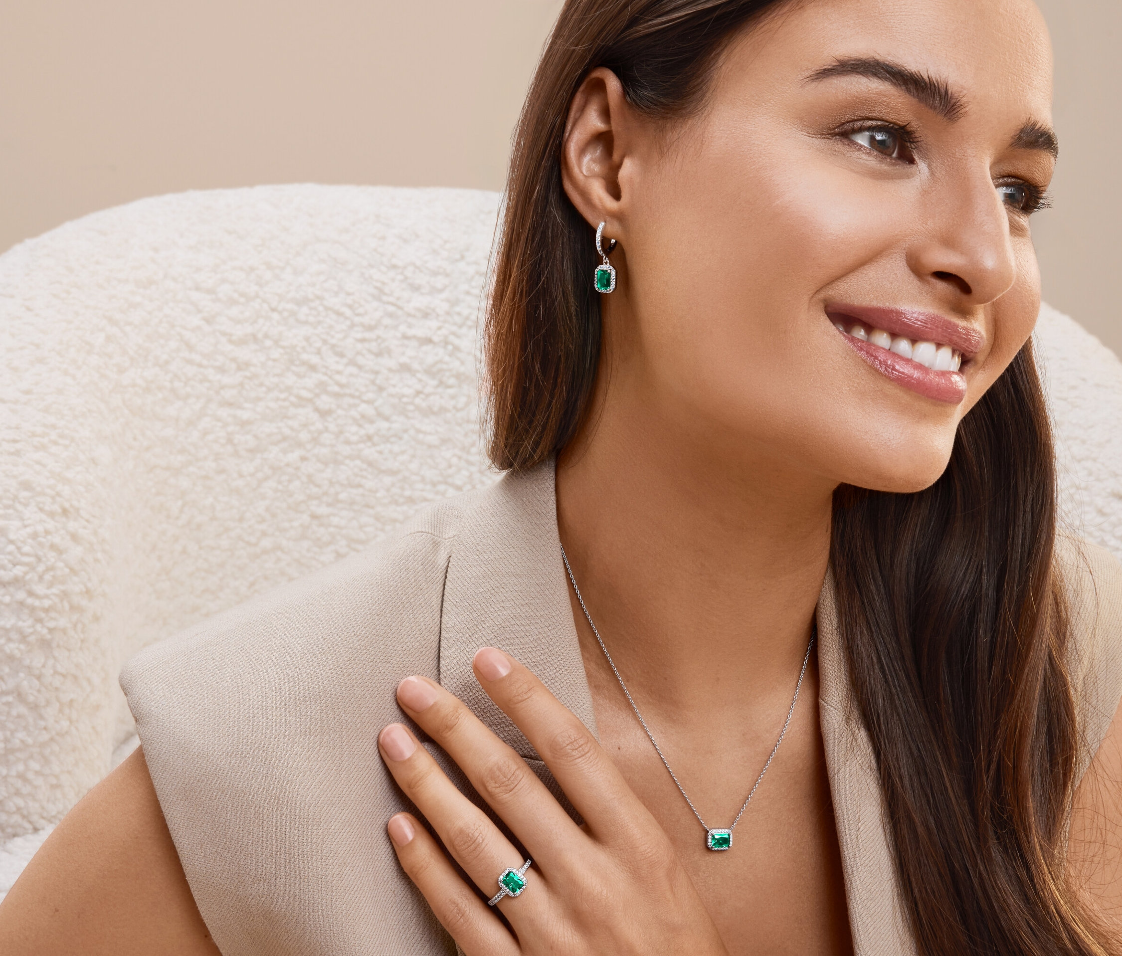 Gülümseyen bir kadın koltukta oturuyor ve zümrüt yeşili taşlı küpeler, kolye ve yüzük takıyor.