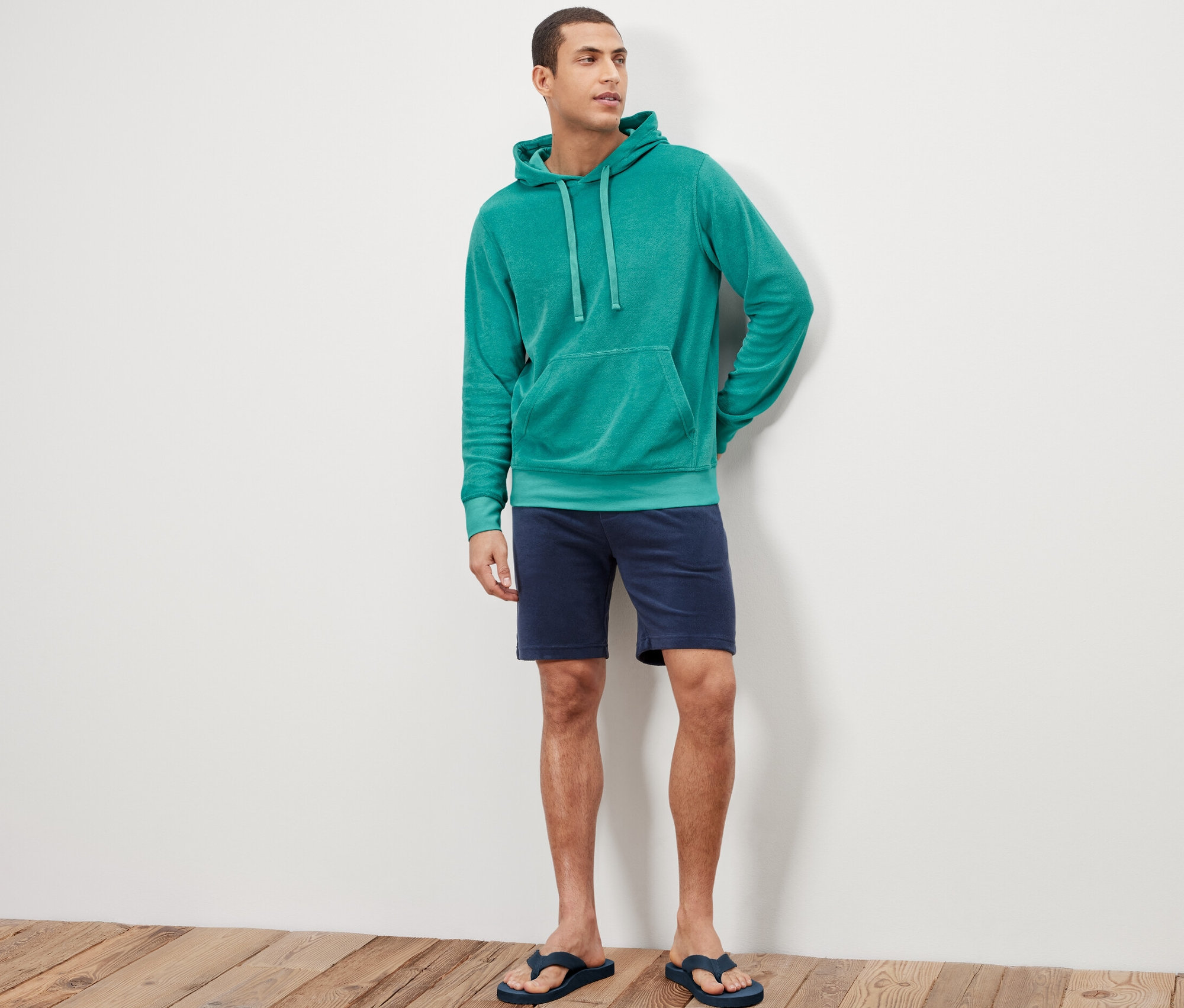 Adam ayakta duruyor ve Havlu Hoodie, şort ve parmak arası terlik giyiyor.