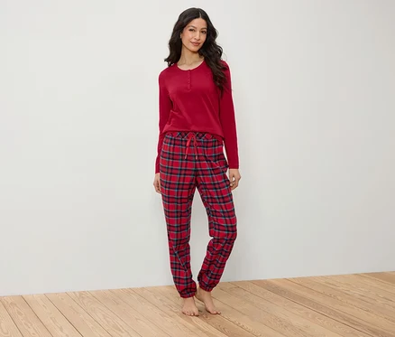 Bir kadın ahşap zeminde duruyor, üzerinde kırmızı uzun kollu bir pijama ve ekose pantolon var.