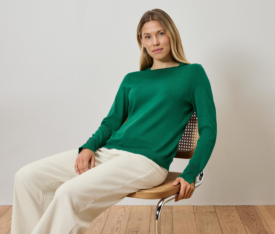 Sarışın bir kadın sandalyede oturuyor, yeşil bir kazak ve krem rengi pantolon giyiyor.