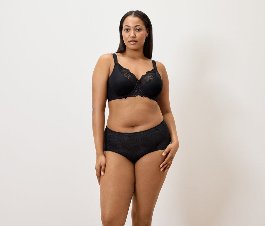 Kadın beyaz bir arka plan önünde siyah toparlayıcı sütyen ve siyah 5'li külot giyiyor.