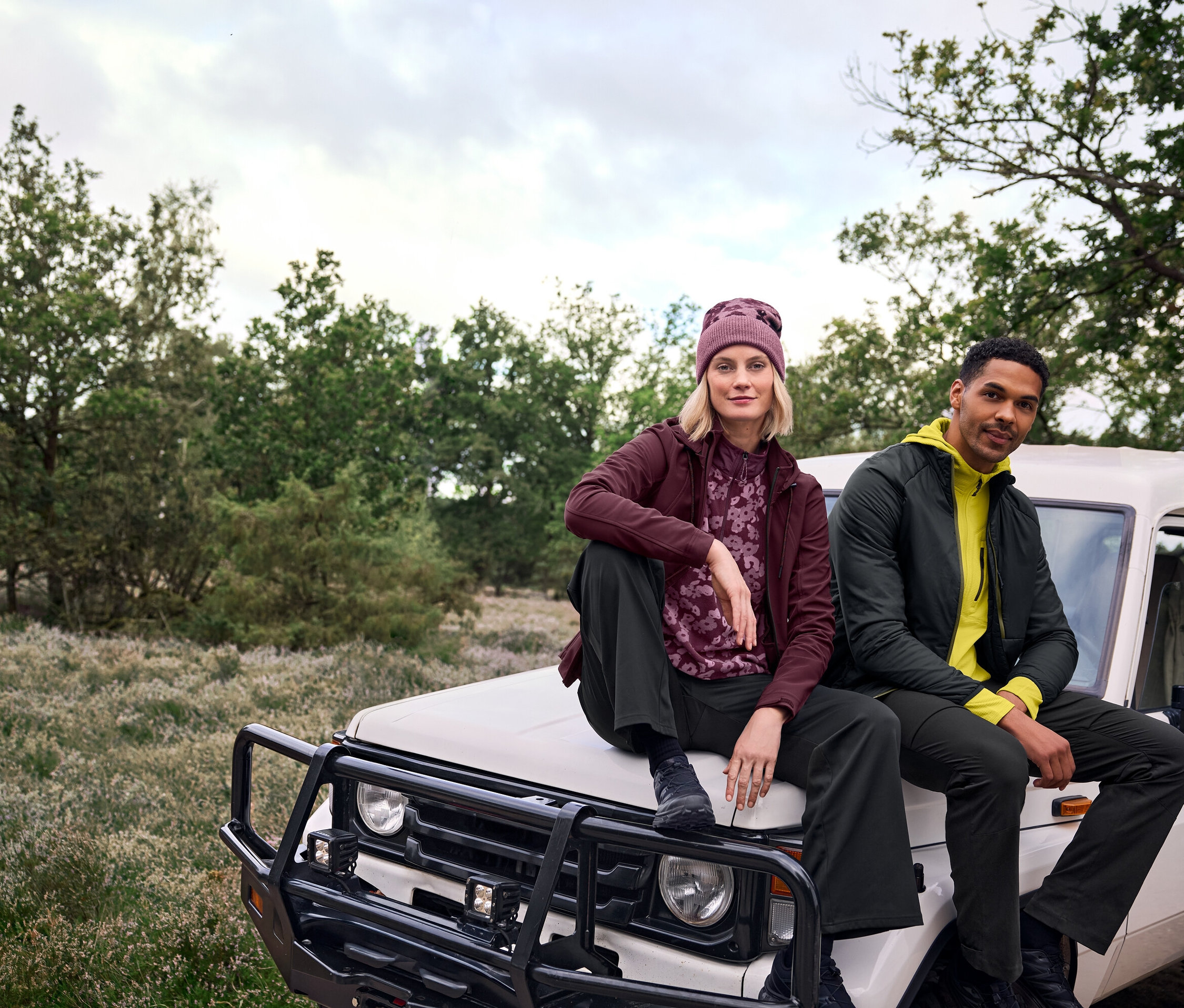 Şapkalı bir kadın ve bir adam doğada beyaz bir arazi aracının kaputunda oturuyor.