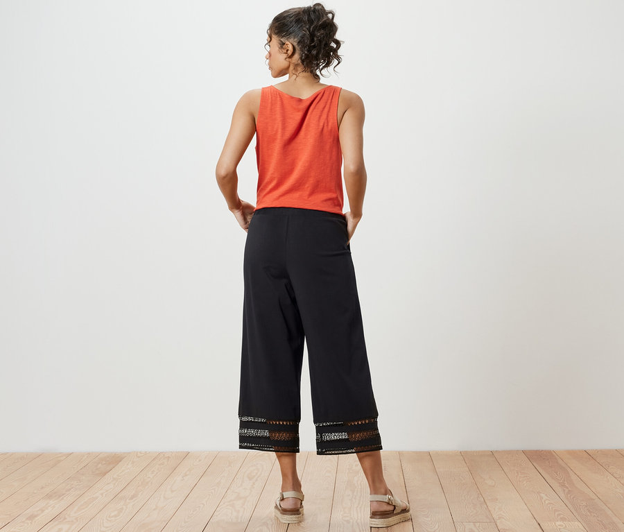 Bir kadın arkası dönük duruyor, üzerinde turuncu bir bluz ve işlemeli siyah culotte pantolon var.