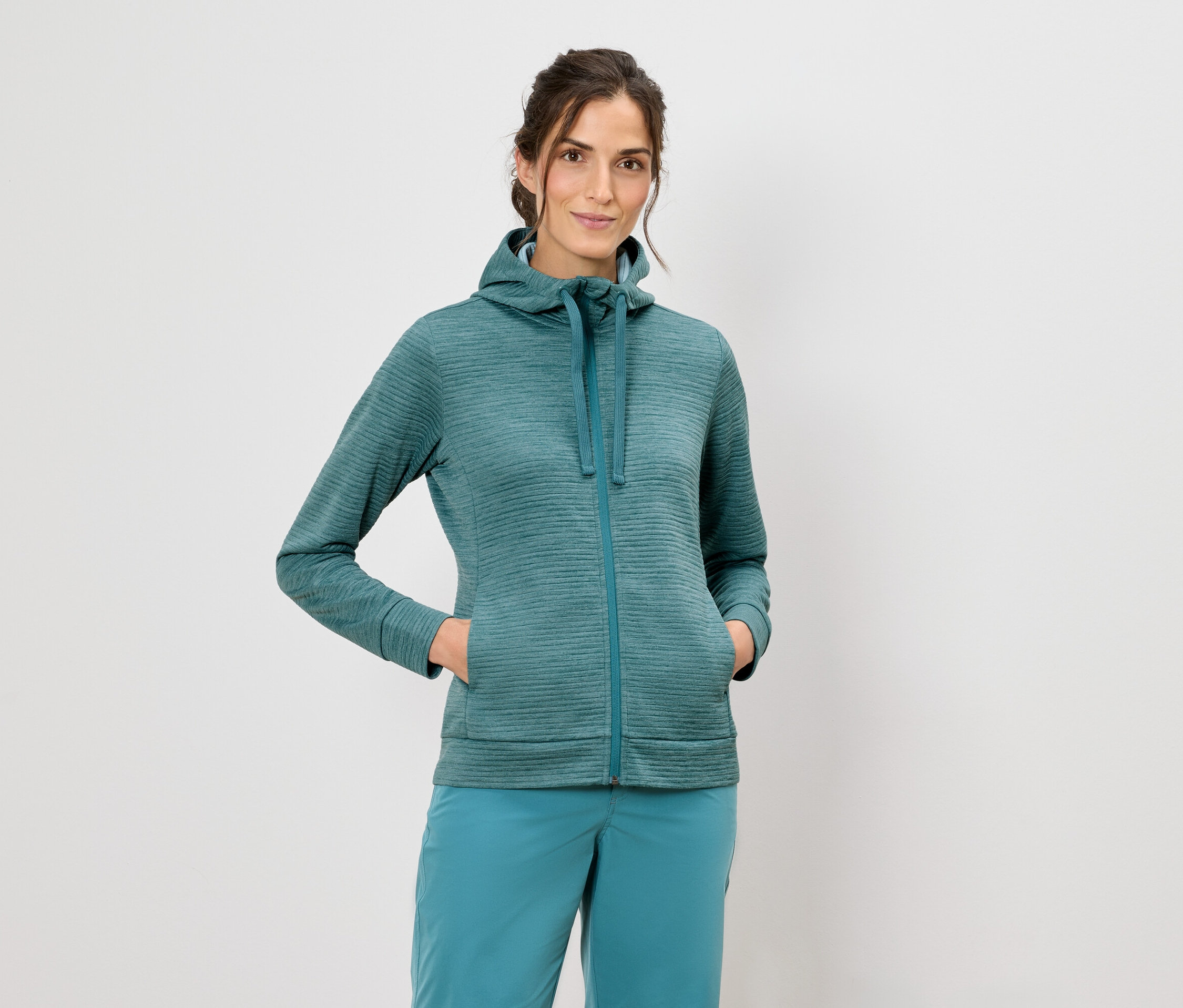 Kapüşonlu ceketli ve mavi pantolonlu bir kadın beyaz bir arka planda duruyor.