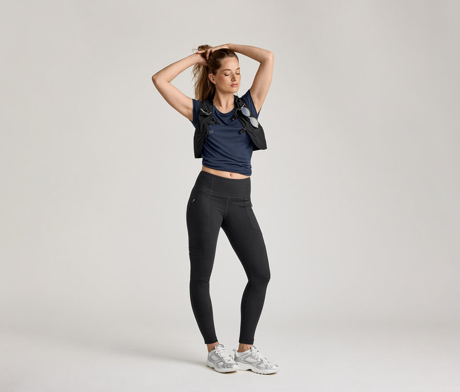 Açık renkli bir arka planda, kadın outdoor tayt, lacivert tişört ve yelek ile poz veriyor.