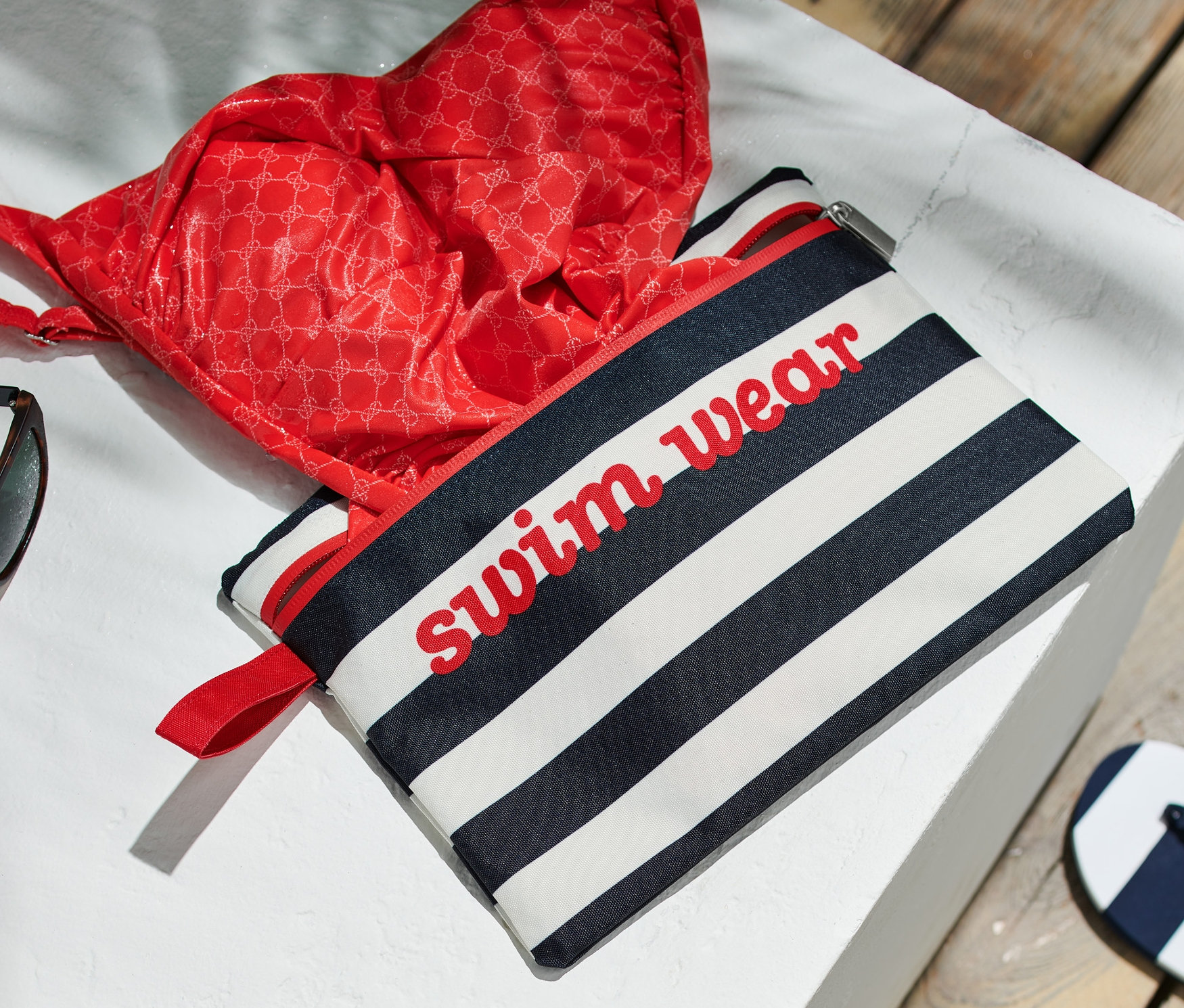 Kırmızı bikini üstü ve güneş gözlüğü, mavi beyaz çizgili Islak Eşya Çantası üzerinde duruyor.