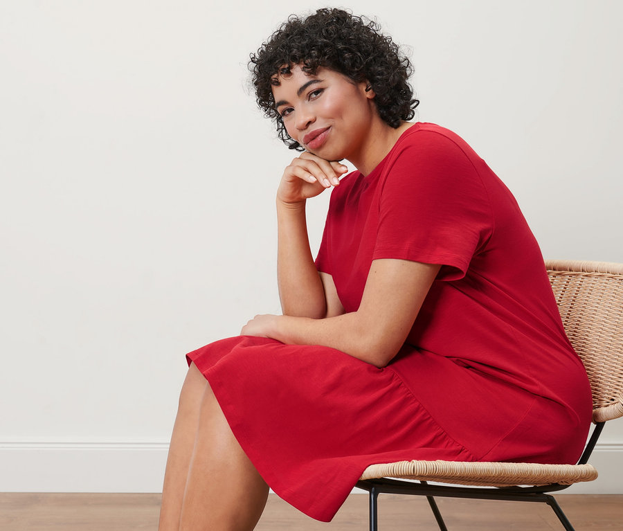 Sandalyede oturan bir kadın kırmızı organik pamuklu volanlı jarse elbise giyiyor.