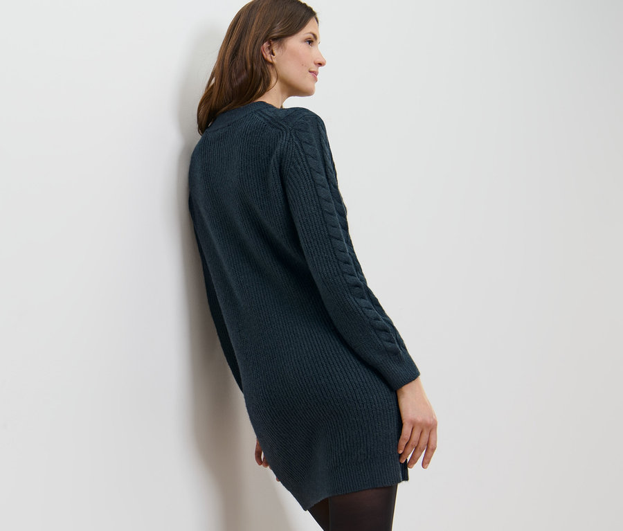 Beyaz duvara yaslanmış motifli örgü elbise giyen bir kadın. 