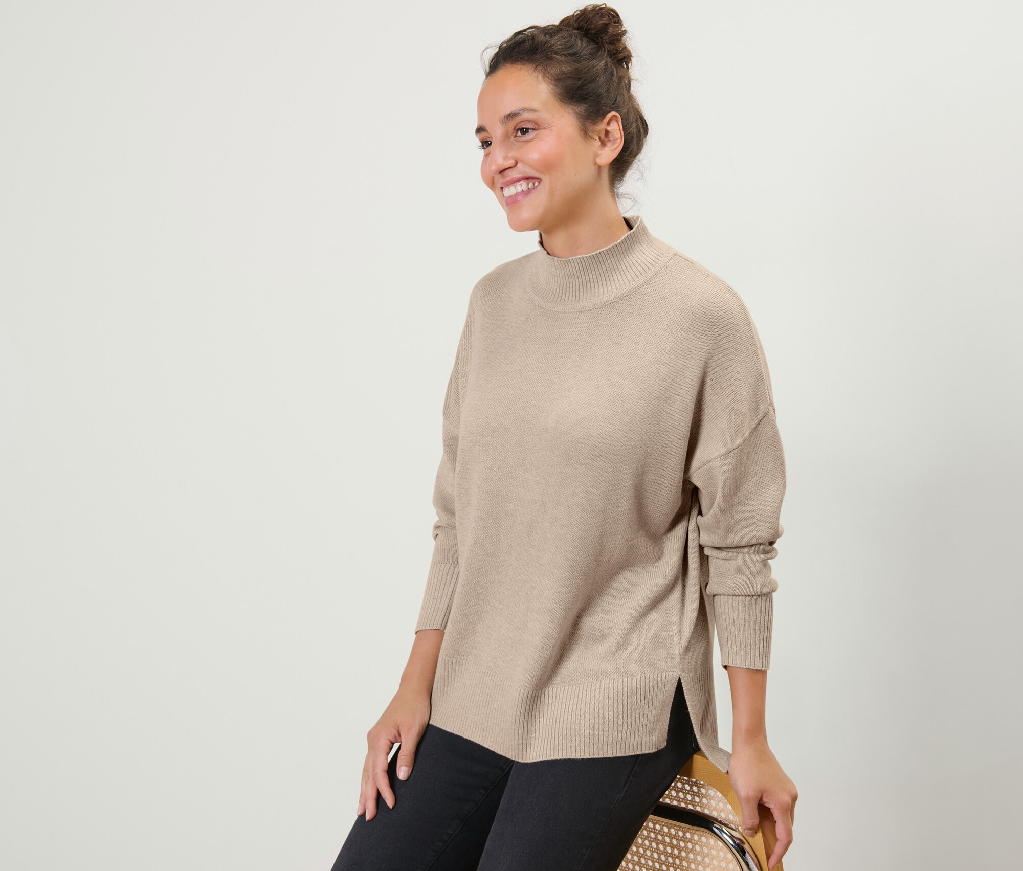 Gülümseyen bir kadın oturuyor, bej rengi yarım balıkçı yaka ince örgü kazak ve siyah kot pantolon giyiyor.